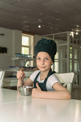 Beautiful young cook chef girl in green chef cap sitting at the table with a spoon and a bowl. Conceptual image of the chefs