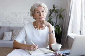Indoor shot of mature woman specialist on retirement working remotely at wooden desk, sitting in...