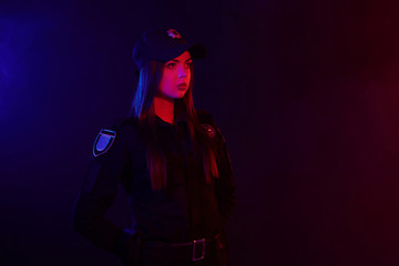Obraz na płótnie Canvas Serious female police officer is posing for the camera against a black background with red and blue backlighting.