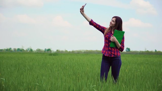 Beautiful young woman student with papers future agronomist makes cute selfie on black mobile in green wheat field. Business meeting male and female on agricultural land
