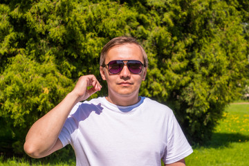 Naklejka na ściany i meble Portrait of a man in sunglasses and a white T-shirt standing outside in park