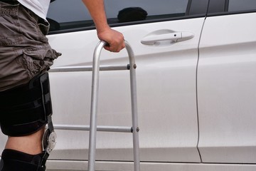 Side view of man wear supporting knee brace and using walker to walk support to white car by himself during rehabilitation therapy