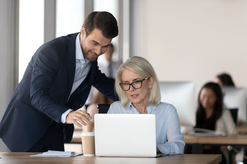 Smiling millennial manager help mature woman with laptop