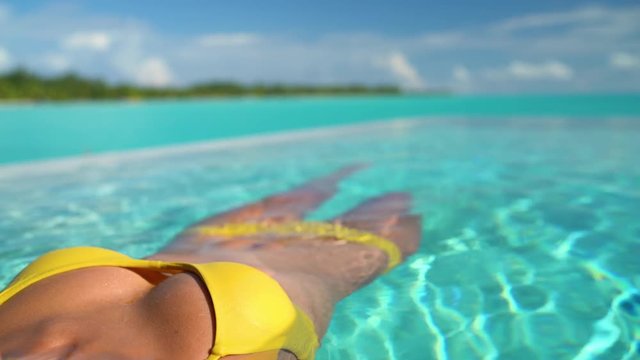 Sexy bikini body woman relaxing swimming in luxury infinity pool of Tahiti resort hotel lying in water floating in yellow bikini. Tanned slim body.