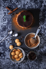 Round tiramisu, homemade no-bake chocolate dessert, traditional Italian sweet food. Table with cake, bowl with cocoa powder, round sponge biscuits,  coffee beans and cinnamon wood. Dark background.