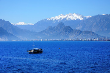 landscape image of Antalya city and high mountains and tour boat on Mediterranean sea over clear sky in Antalya, Turkey