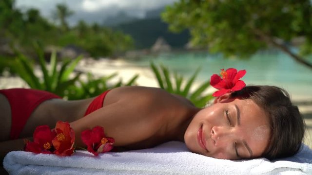 Woman relaxing at spa luxury massage hotel resort. Beauty girl lying down sleeping on towel with hibicus flowers outside. Serene ethnic woman relaxing. Mixed race Asian / Caucasian model outdoor.
