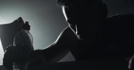 Young tired man drinking from the water bottle after training in the dark boxing ring with smoke. Silhouette. Boxing concept. - Powered by Adobe