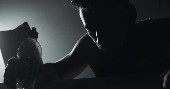 Young tired man drinking from the water bottle after training in the dark boxing ring with smoke. Silhouette. Boxing concept.
