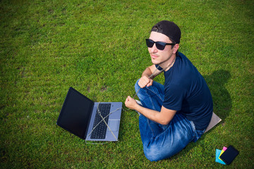 A young man with a chain around his neck is tied to a laptop sitting on the grass with a vignetting effect. The concept of freelancing and self-organization, commitment and affection