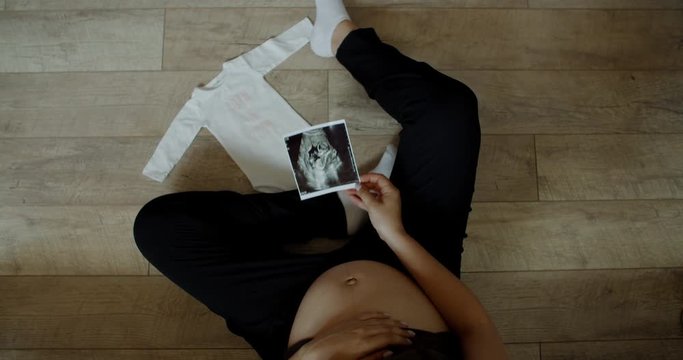 Caucasian pregnant women sitting on floor near bed with babies clothes and picking up sonogram image. 60 fps