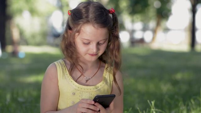 A girl sits on grass in a park on a sunny summer day and communicates with friends in social networks using a smartphone. Smiling  teenager chatting using mobile phone, writes letter, sms. Browse phot