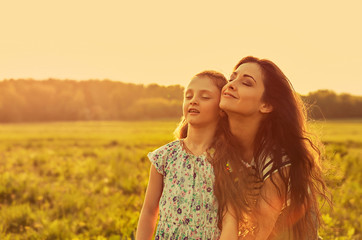 Happy enjoying mother hugging her relaxing joying kid girl and breathing fresh air with closed eyes on sunset bright summer background. Closeup
