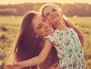 Happy enjoying mother hugging her relaxing joying kid girl on sunset bright summer background. Closeup portrait