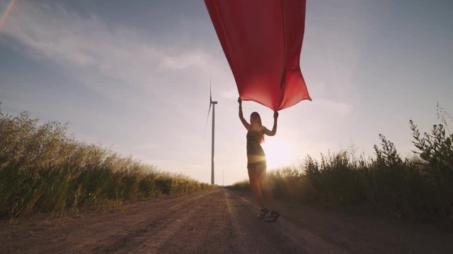 Woman with pieces of red cloth dance near the field with wind generators . Windmill on sunset. Slow motion. Stylish hipster woman