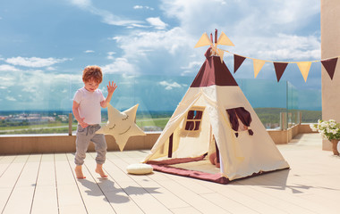 happy young boy, kid playing near the textile wigwam tent on the summer patio © Olesia Bilkei