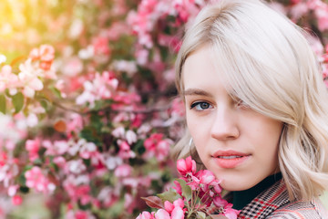 Fototapeta premium Close up portrait of young pretty caucasian curly blondie model posing outdoors in pink blossom garden. Beauty, nature concept
