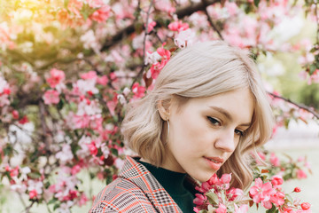 Fototapeta premium Close up portrait of young pretty caucasian curly blondie model posing outdoors in pink blossom garden. Beauty, nature concept