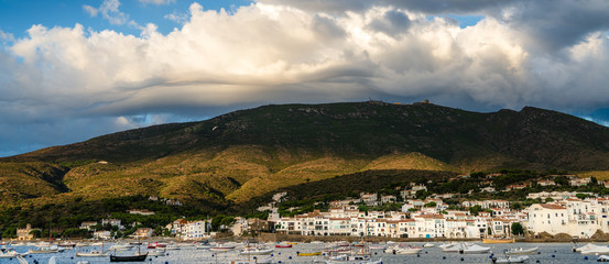 Dramatic clouds form over the hills above the bay and seaside town of Cadaques, Girona, Catalonia, Spain. Boats moored on the water and sun light hitting the hills.