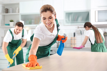 Woman using rag and sprayer for cleaning table with colleagues in kitchen