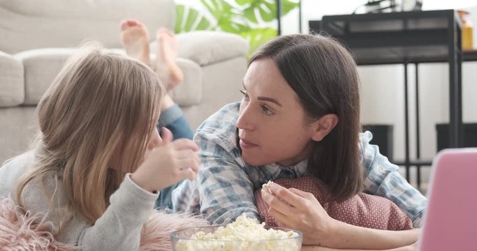 Mother and little daughter discussing while eating popcorn and watching movie on laptop