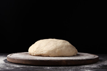 Wooden board with flour and bread dough on table against dark background. Space for text