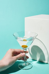 partial view of woman holding transparent glass with cocktail and olive near white cube on turquoise background