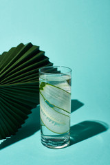 transparent glass with fresh water and cucumber slices near green leaf on turquoise background