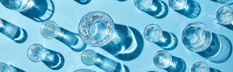 top view of transparent glasses with clear water and shadows on blue background, panoramic shot