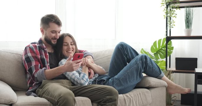 Happy young couple using mobile phone and talking with each other while lying on sofa at home