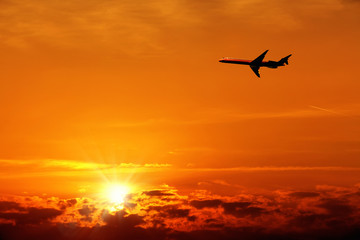 Fototapeta na wymiar Silhouetted flying airplane over beautiful cloudscape of orange colored sunset sky with shining sun