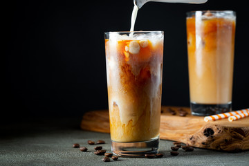 Ice coffee in a tall glass with cream poured over, ice cubes and beans on a old rustic wooden table. Cold summer drink with tubes on a black background with copy space