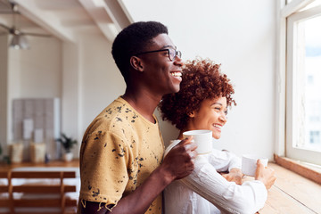 Young Couple Relaxing In Loft Apartment Looking Out Of Window With Hot Drink