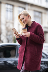 Young woman standing at the street drinking coffee to go and using mobile phone