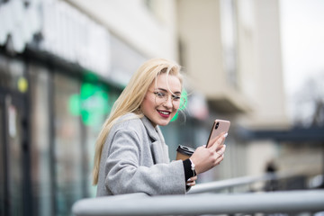 Fototapeta premium Street portrait of young smiling blonde woman wearing grey coat using phone
