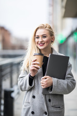 Fototapeta premium Portrait of smiling woman strolling through city street with silver laptop and takeaway coffee in hands