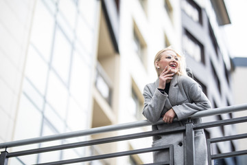 Happy fashion woman in gray coat leaning on handrail in city street.