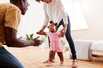 Parents At Home Encouraging Baby Daughter To Take First Steps
