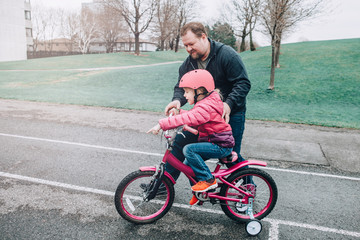 Caucasian father dad training helping girl daughter to ride bicycle. Preschooler child kid in pink helmet with bike on backyard road outside on spring day. Seasonal child family outdoor activity