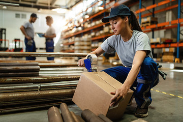Young woman working in warehouse and taping cardboard box for shipment.