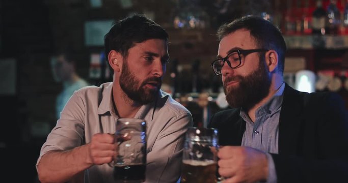 Two old friends drinking draft beer and talking in the sport bar. Friends sitting at bar counter, drinking beer and talking in beer pub.