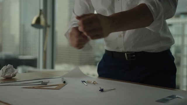 Businessman rolls up sleeves and starts to work on blueprint in office with view on downtown and skyscrapers. Architect's work process.