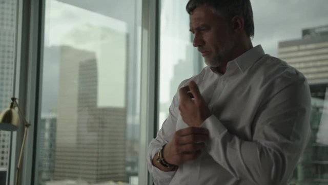 Businessman rolls up sleeves and starts to work on blueprint in office with view on downtown and skyscrapers. Architect's work process.