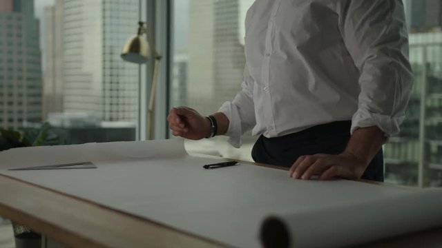 Businessman rolls up sleeves and starts to work on blueprint in office with view on downtown and skyscrapers. Architect's work process.