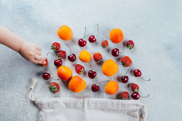 Various fresh summer berries - strawberry, cherry and apricot. Baby hand takes berry. Reusable bag, tote bag, minimal waste. Top view, flat lay