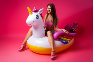 Concept summer mood, relaxation and beauty.Modern portrait of a young woman in a shorts, top and sandals resting on an inflatable unicorn mattress on an isolated pink background © Виталий Сова