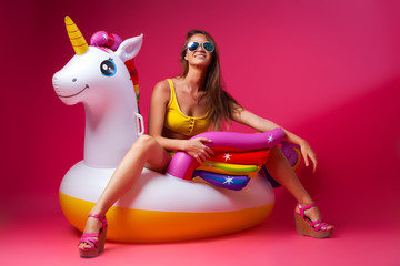 Concept summer mood, relaxation and beauty.Modern portrait of a young woman in a shorts, top and sandals resting on an inflatable unicorn mattress on an isolated pink background © Виталий Сова