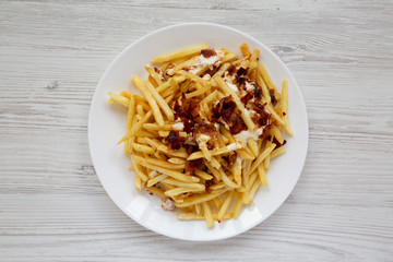 French fries with cheese sauce and bacon on a white plate over white wooden background, overhead view. Flat lay, from above, top view.