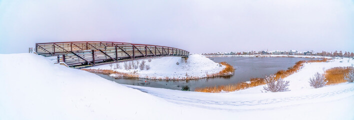 Panorama of a silvery lake with snow covered shore and metal bridge