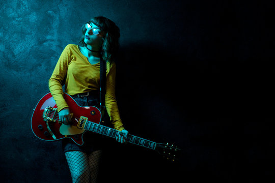 Sensual young hipster woman with curly hair with red guitar in neon lights. Rock musician is playing electrical guitar. 90s style concept.